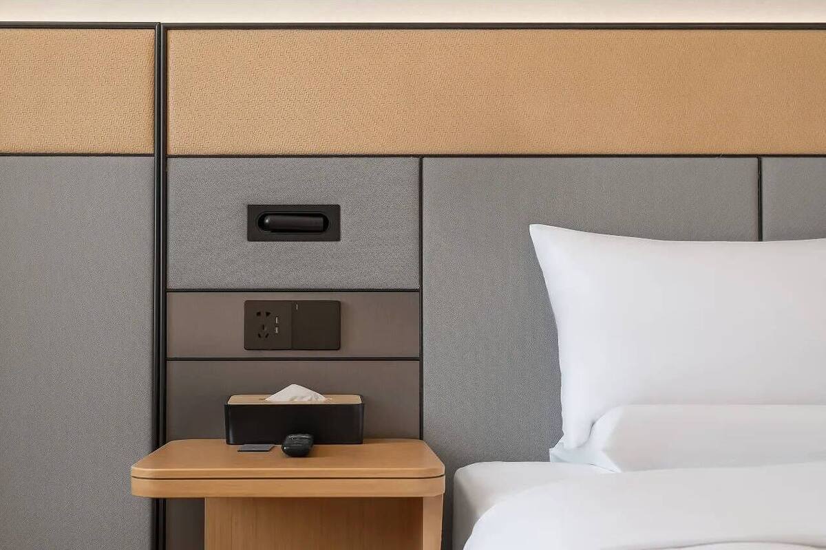 Habitación de hotel con cama, mesita de noche con caja de pañuelos y control remoto, y panel de pared con toma de corriente e interruptor.