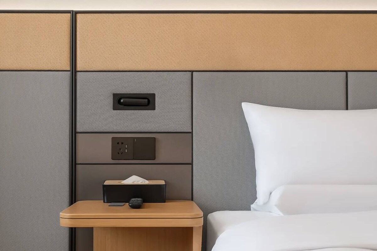 A hotel room with a bed, a nightstand with a tissue box and remote control, and a wall panel with a power outlet and switch.