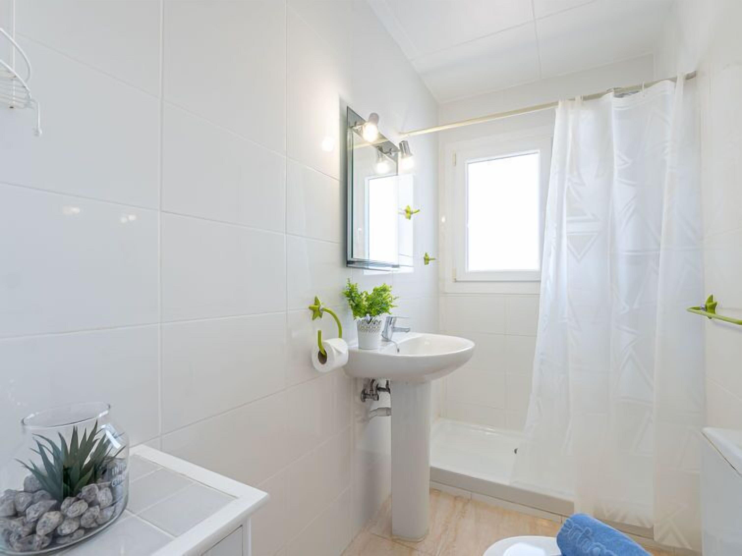 A modern bathroom with white tiles, a sink, a mirror, a shower with a curtain, and a small plant on the counter.