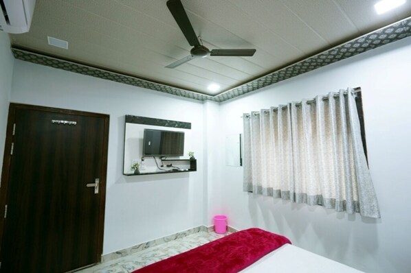 Un dormitorio con ventilador de techo, una puerta, un espejo, una ventana con cortinas y un cubo de basura rosa.