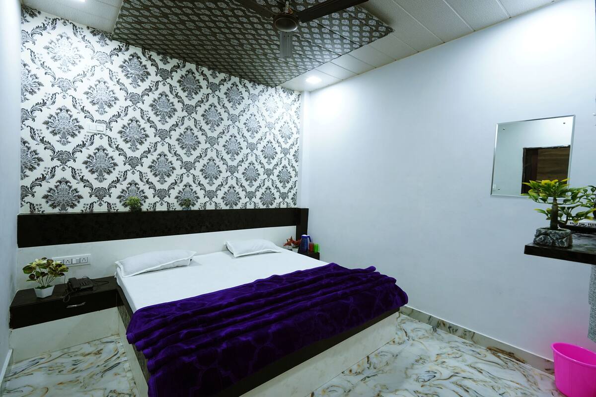 Un dormitorio con papel tapiz estampado, una cama con manta morada, una mesita de noche con un teléfono y una planta, y un pequeño estante con una planta.