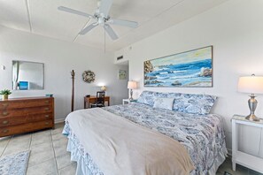 A bedroom with a bed, dresser, desk, chair, lamps, and a painting of a beach scene.