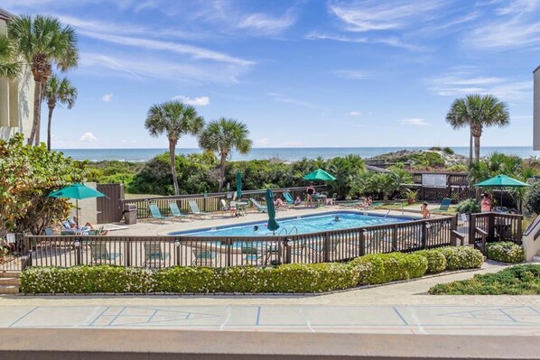 A pool area with lounge chairs and umbrellas, surrounded by palm trees and a view of the ocean.