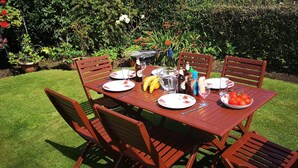 Mesa de madera al aire libre con sillas, platos, cubiertos, un bol de tomates, una jarra y botellas.