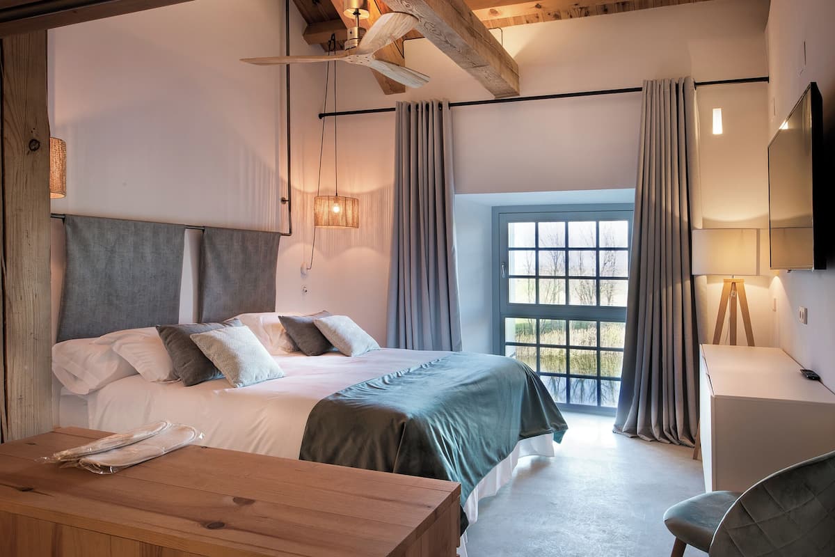 Un dormitorio moderno con techo de madera, una cama grande con edredón gris y blanco, una mesita de noche de madera, un escritorio, una silla y una ventana con cortinas.