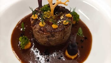 Un plato con un bistec en el centro, adornado con hierbas verdes y una estructura decorativa que parece un árbol, rodeado de salsa y pequeñas guarniciones.