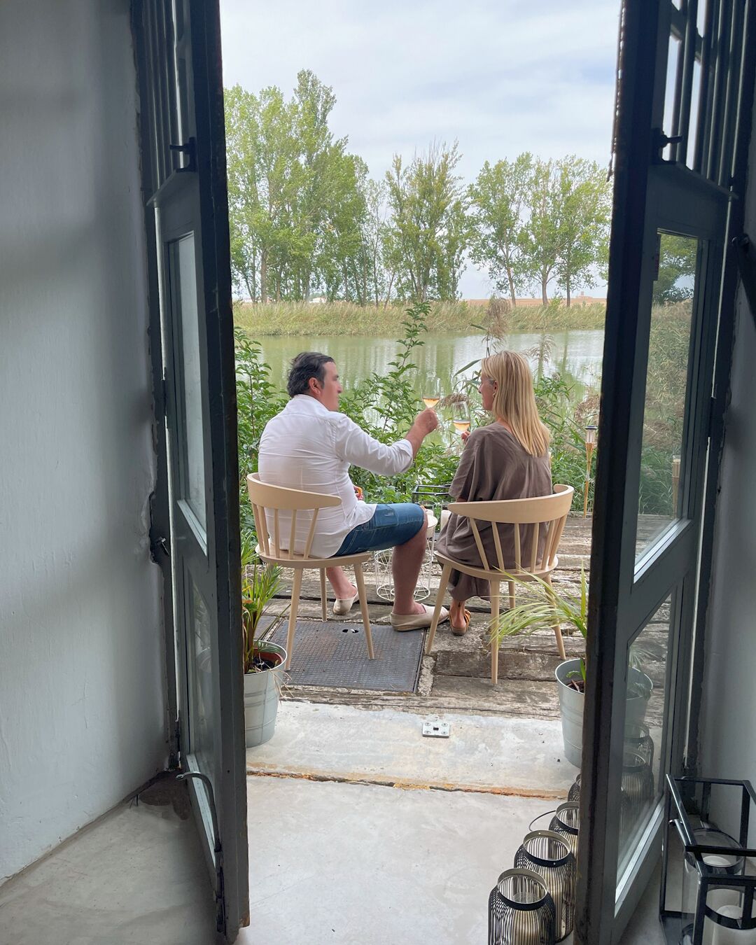 Deux personnes sont assises à une table près d’une fenêtre donnant sur un plan d’eau. On peut voir des chaises et des plantes en pot.