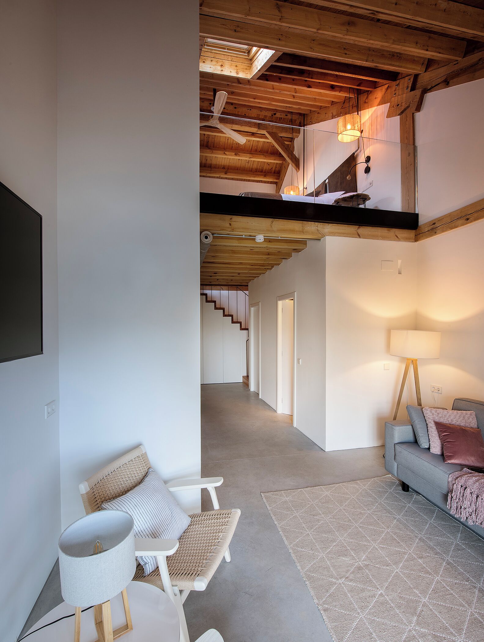 A modern living room with a high ceiling, exposed wooden beams, a sofa set, a floor lamp, and a flat-screen TV mounted on the wall.