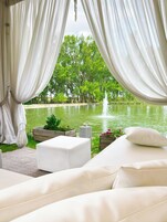 Un espacio exterior tranquilo con cortinas blancas, un sofá blanco y vistas a un estanque con una fuente.