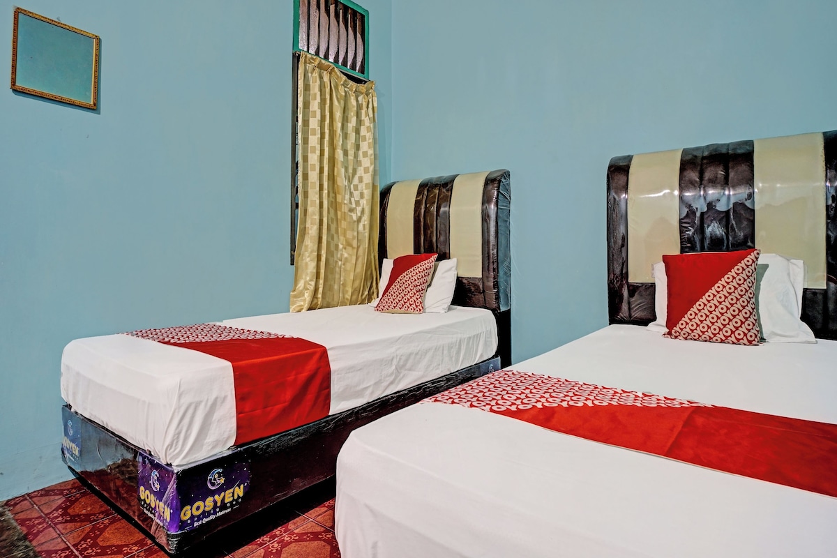 A room with two beds, each with a white mattress and red bedding. There is a window with a curtain, and a framed picture on the wall.