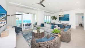 A spacious living room with a large TV, comfortable seating, a ceiling fan, and a view of the ocean through large windows. - Rum Point Resort 201 by Grand Cayman Villas (Seven Mile Beach)