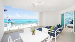 Quarto moderno com mesa e cadeiras brancas, sofá azul com almofadas verdes e vista para o oceano através de grandes janelas.