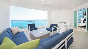 A modern living room with a large sofa, armchairs, and a coffee table. It features a balcony with a view of the ocean. - Rum Point Resort 405 by Grand Cayman Villas (Seven Mile Beach)