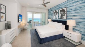 A modern hotel room with a bed, bedside table, desk, and chair. There is a view of the ocean through large windows. - Rum Point Resort 405 by Grand Cayman Villas (Seven Mile Beach)