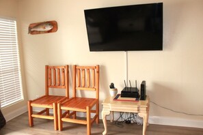 Et værelse med to træstole, et lille bord med en router og en bog, samt et fladskærms-tv monteret på væggen.