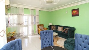 A living room with a green wall, a green sofa, a blue armchair, a wooden chair, and a small round table. - Furnished apartment in Douala, Makepe (Douala)