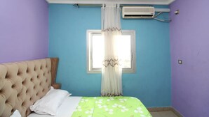 A bedroom with a tufted headboard, a bed with a green floral bedspread, a window with white curtains, and an air conditioning unit on the wall. - Furnished apartment in Douala, Makepe (Douala)