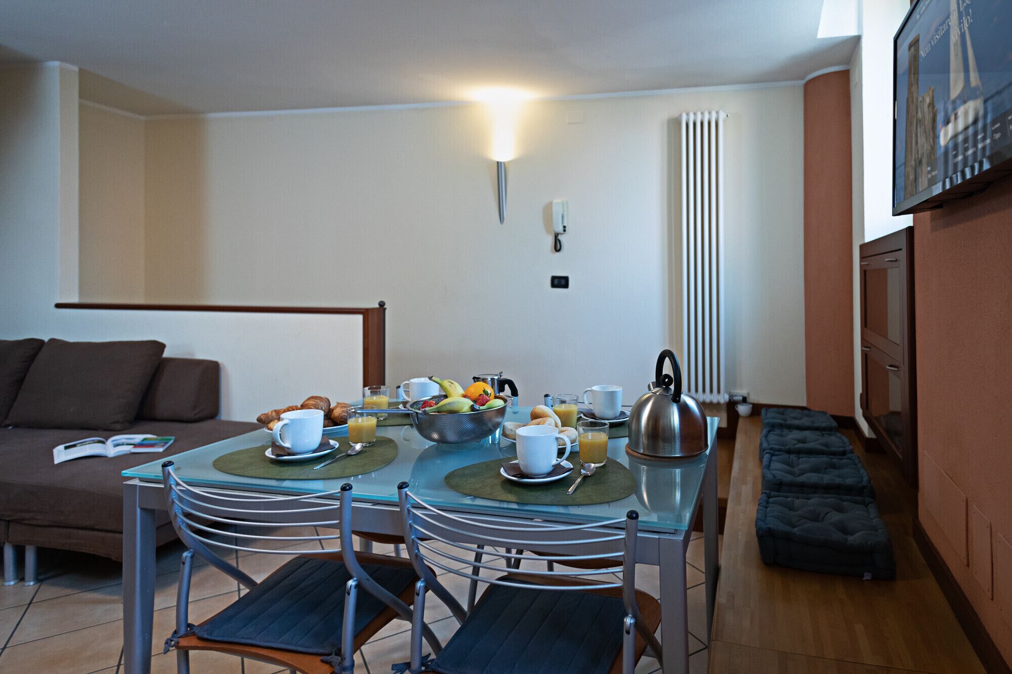 Uma área de refeições com uma mesa posta para o pequeno-almoço, incluindo croissants, fruta e bebidas.