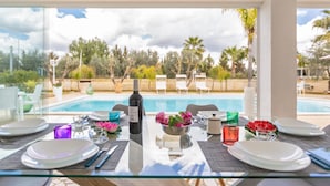 A dining table set for two with a view of a swimming pool and palm trees. - 3220 Villa Smeralda by Barbarhouse (Guagnano)