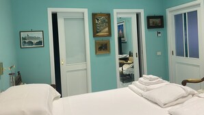 A room with teal walls, white beds, and doors leading to other rooms. There are framed pictures on the walls and a bed with neatly folded white towels.