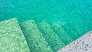 Une piscine aux eaux cristallines, accessible par des marches, entourée d’un mur de pierre et offrant une vue sur l’océan et les collines.