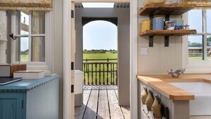 Una habitación con vistas a un campo verde a través de una puerta abierta, una terraza de madera y una cocina con un fregadero y estanterías.