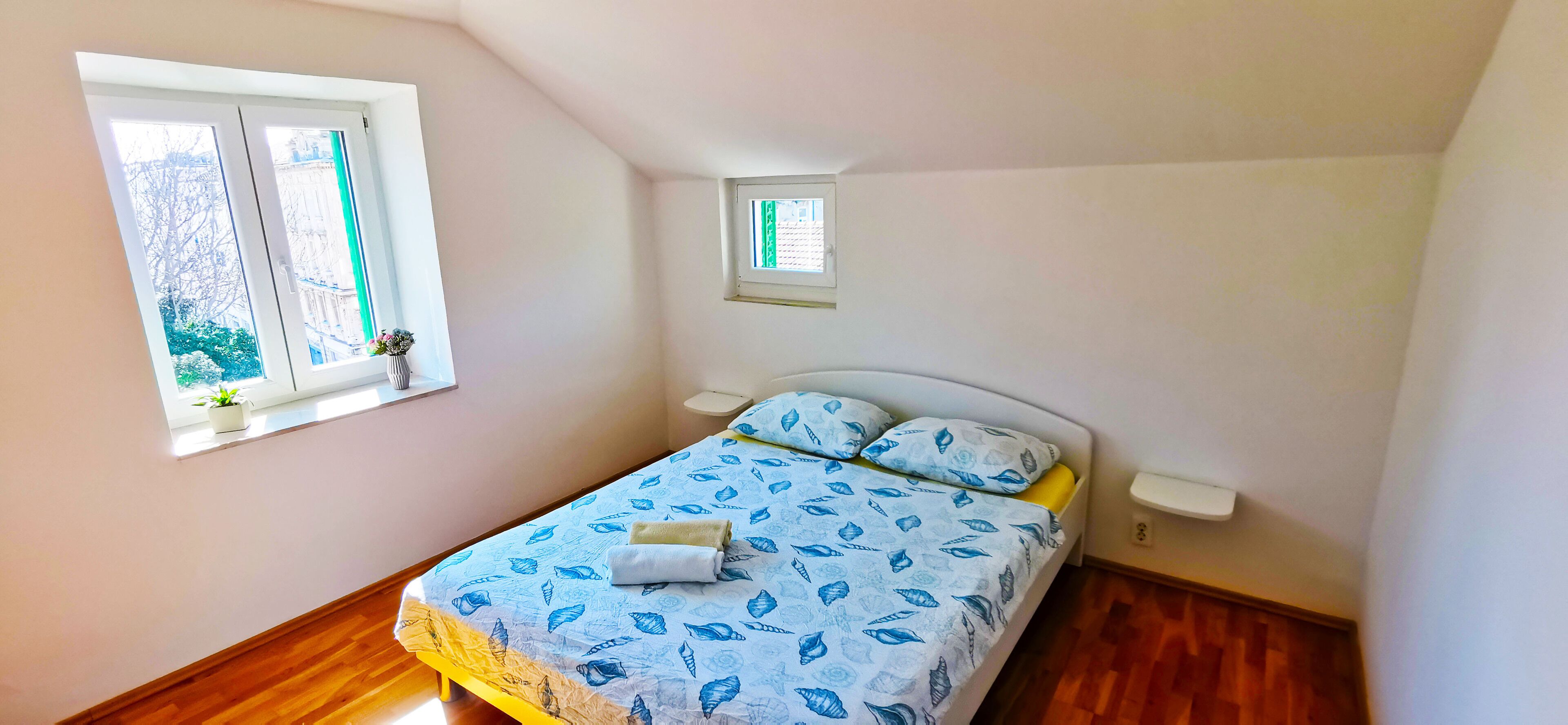 Un dormitorio con una cama, dos ventanas, una mesita y una mesilla de noche.