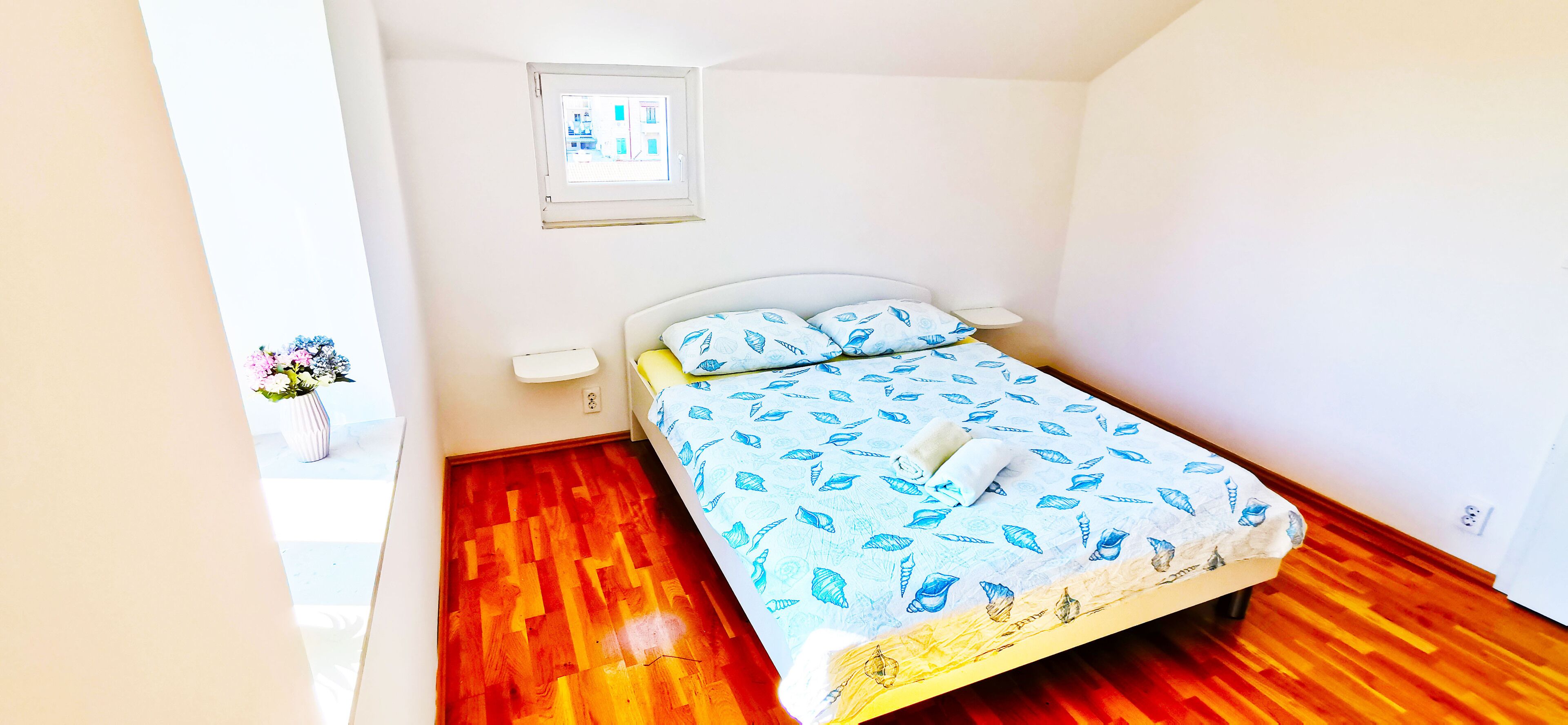 A bedroom with a bed, a nightstand, a window, and a wall-mounted shelf with flowers.