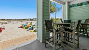 Une terrasse couverte, dotée de chaises de plage et d’une table, qui offre une vue sur une plage de sable où l’on aperçoit des chaises colorées et un palmier.