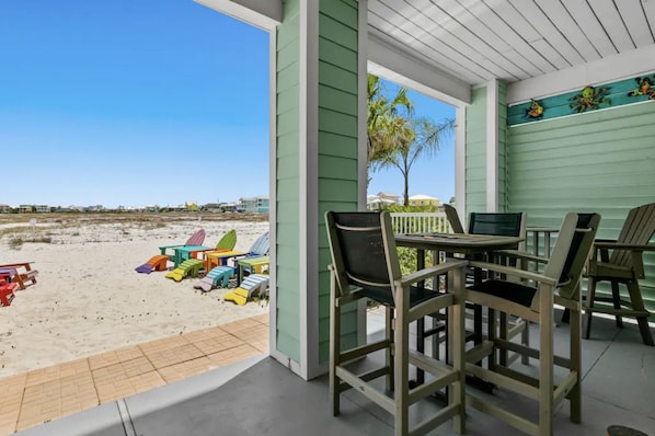 Eine überdachte Terrasse mit Liegestühlen und einem Tisch, mit Blick auf einen Sandstrand mit bunten Stühlen und einer Palme.
