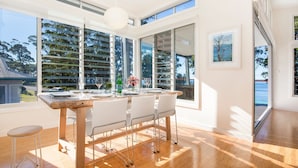 Een eetruimte met een houten tafel, witte stoelen en een glazen balustrade met uitzicht op een waterpartij.