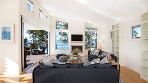 A spacious living room with a fireplace, large windows, and a view of the outdoors. - Hyams Drift + Studio (Hyams Beach)