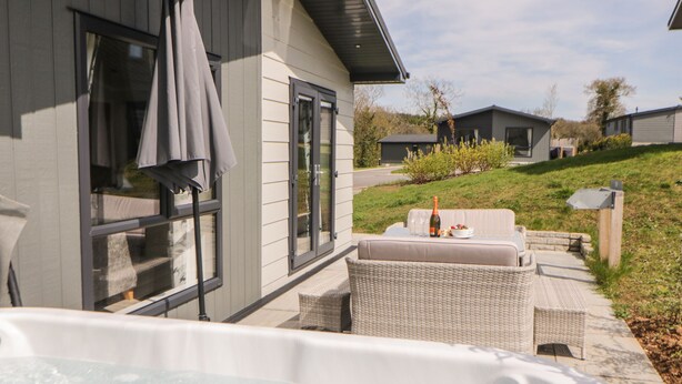 A hot tub in front of a house with a patio, a bottle of champagne, and a bowl of fruit on the table. - Meadow Lodge (Lifton)