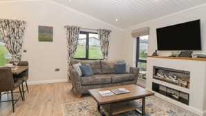 A spacious living room with a sofa, coffee table, and fireplace. There is a dining area with a table and chairs, and a large window with curtains. - Meadow Lodge (Lifton)