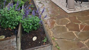 Un jardín con jardineras elevadas y un patio con suelo de piedra y mobiliario exterior.