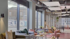 Un interior moderno con amplios ventanales, un área de descanso con sofás y mesas, y detalles arquitectónicos visibles como conductos expuestos e iluminación de techo.