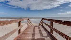 Un muelle de madera que conduce a una playa, con cielo despejado y el océano al fondo.