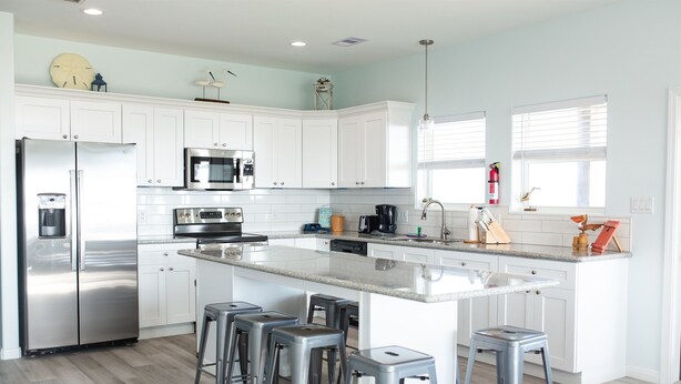 Una cocina moderna con armarios blancos, electrodomésticos de acero inoxidable, una isla grande con taburetes y una ventana con persianas.