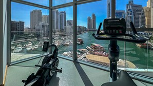 Vue depuis une chambre où se trouve un vélo d’appartement, donnant sur une marina avec des bateaux et sur un paysage urbain avec de hauts immeubles.