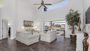 Ein geräumiges Wohnzimmer mit gestreiften Sofas, einem Deckenventilator und einem großen Fenster mit Blick auf einen Pool.