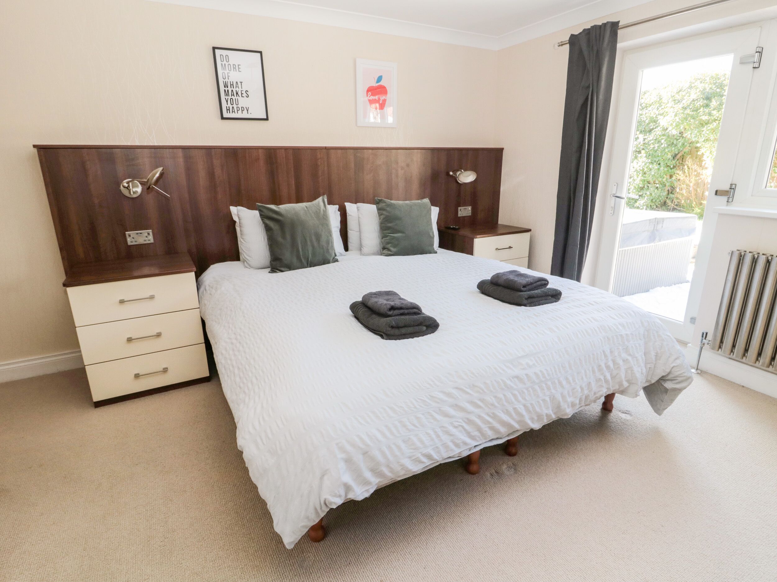 A neatly made bed with a white duvet and grey towels, a wooden headboard, a bedside table with a lamp, and a small drawer unit.
