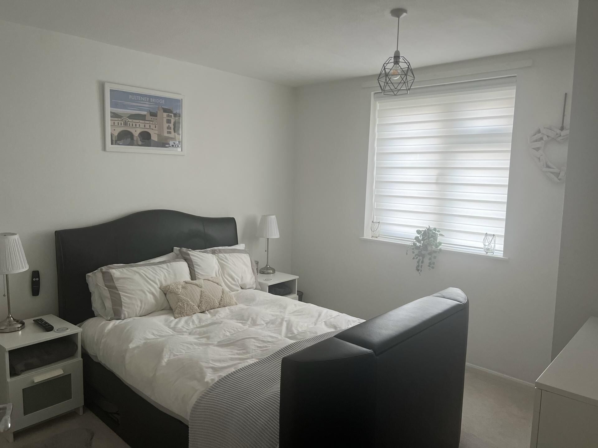 A bedroom with a bed, bedside tables, a lamp, a hanging plant, and a window with blinds.