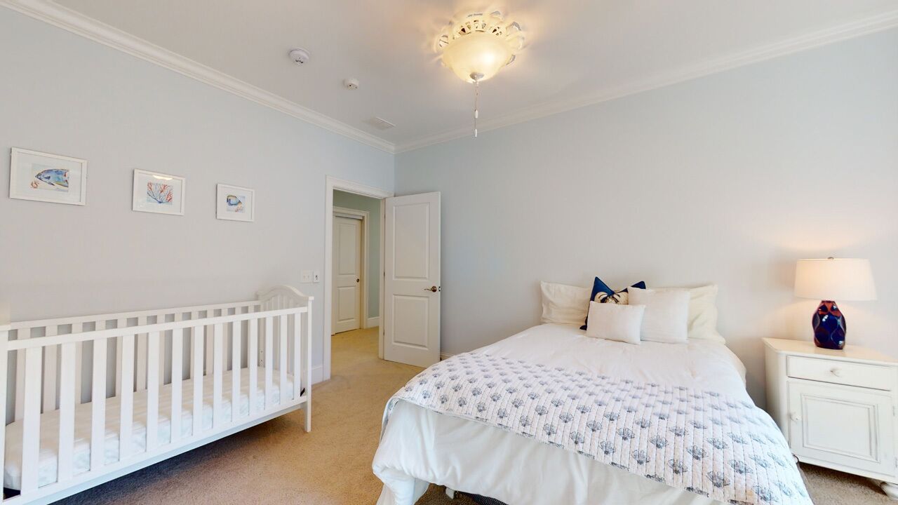A bedroom with a crib, a bed with a patterned bedspread, a nightstand with a lamp, and framed pictures on the wall.