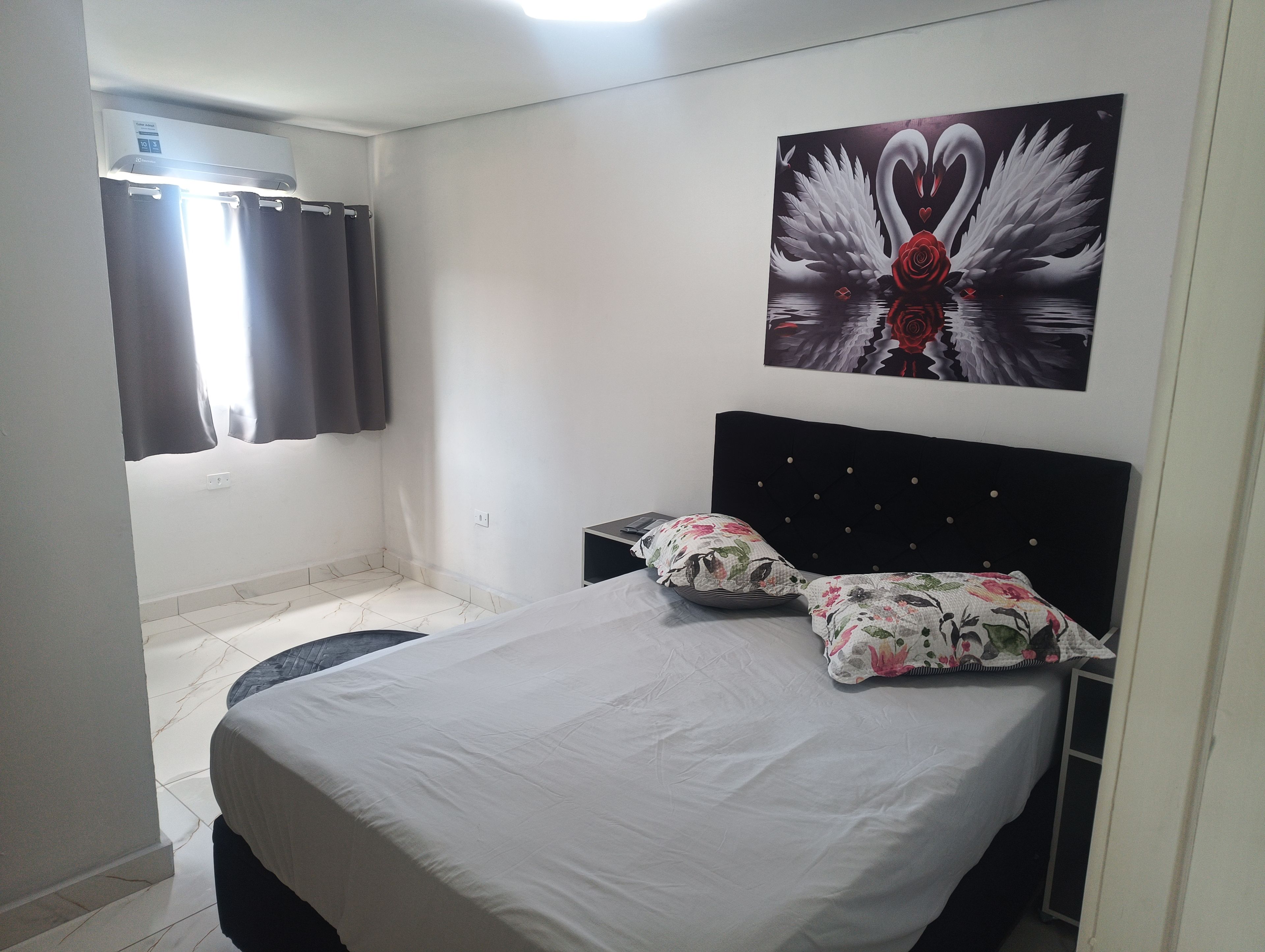 Quarto com cama, mesa de cabeceira, ar condicionado fixado na parede e janela com cortinas.