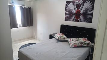 Quarto com cama, mesa de cabeceira, ar condicionado fixado na parede e janela com cortinas.