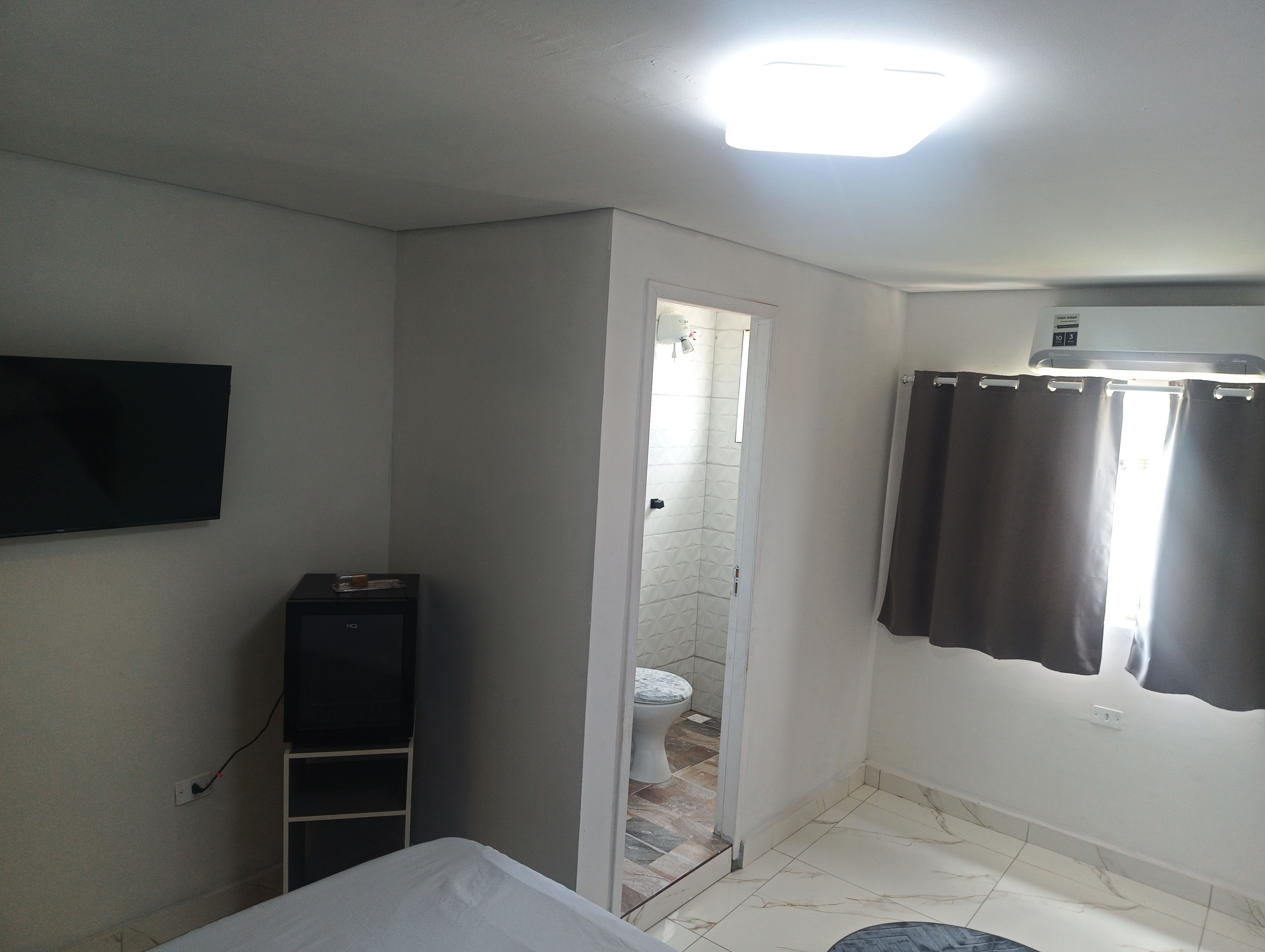 A hotel room with a bed, a flat-screen TV mounted on the wall, a small cabinet, a shower area, and a curtain.