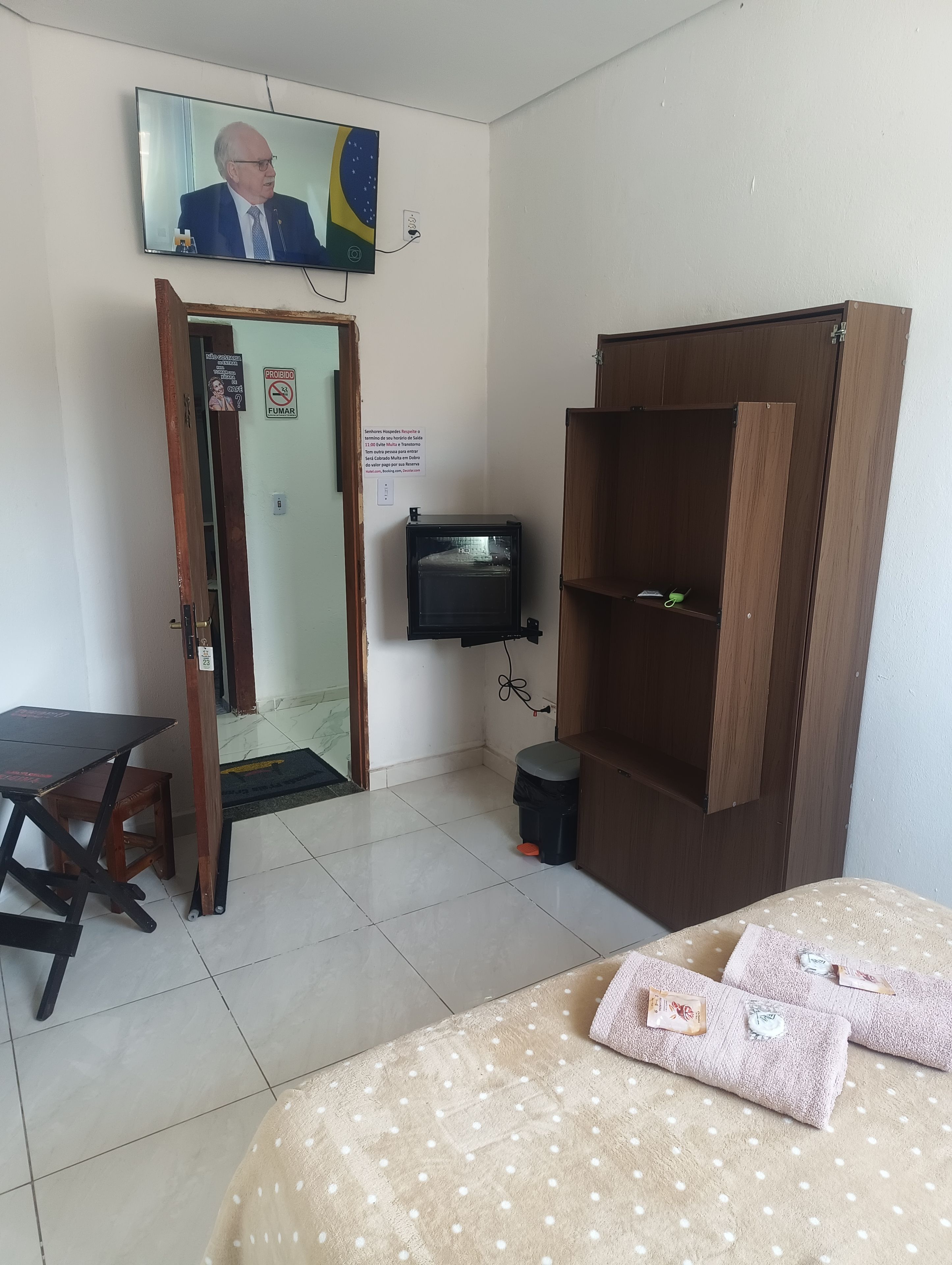 Une chambre avec un lit, une armoire en bois, un téléviseur fixé au mur et une petite table avec une chaise.