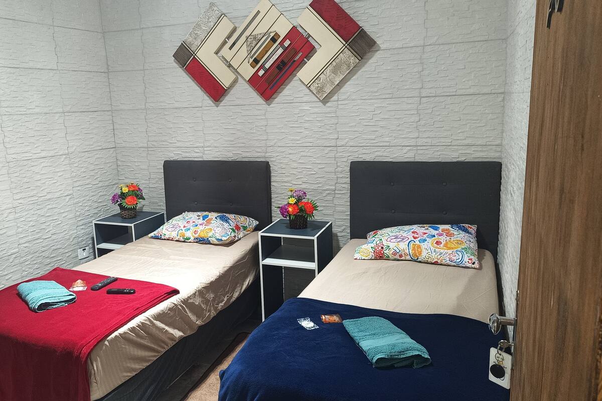 A room with two beds, each with a different colored blanket, a small table with a vase of flowers, and a wall-mounted artwork.