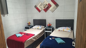 A room with two beds, each with a different colored blanket, a small table with a vase of flowers, and a wall-mounted artwork. - Pousada Praia Grande Solemar (Praia Grande)