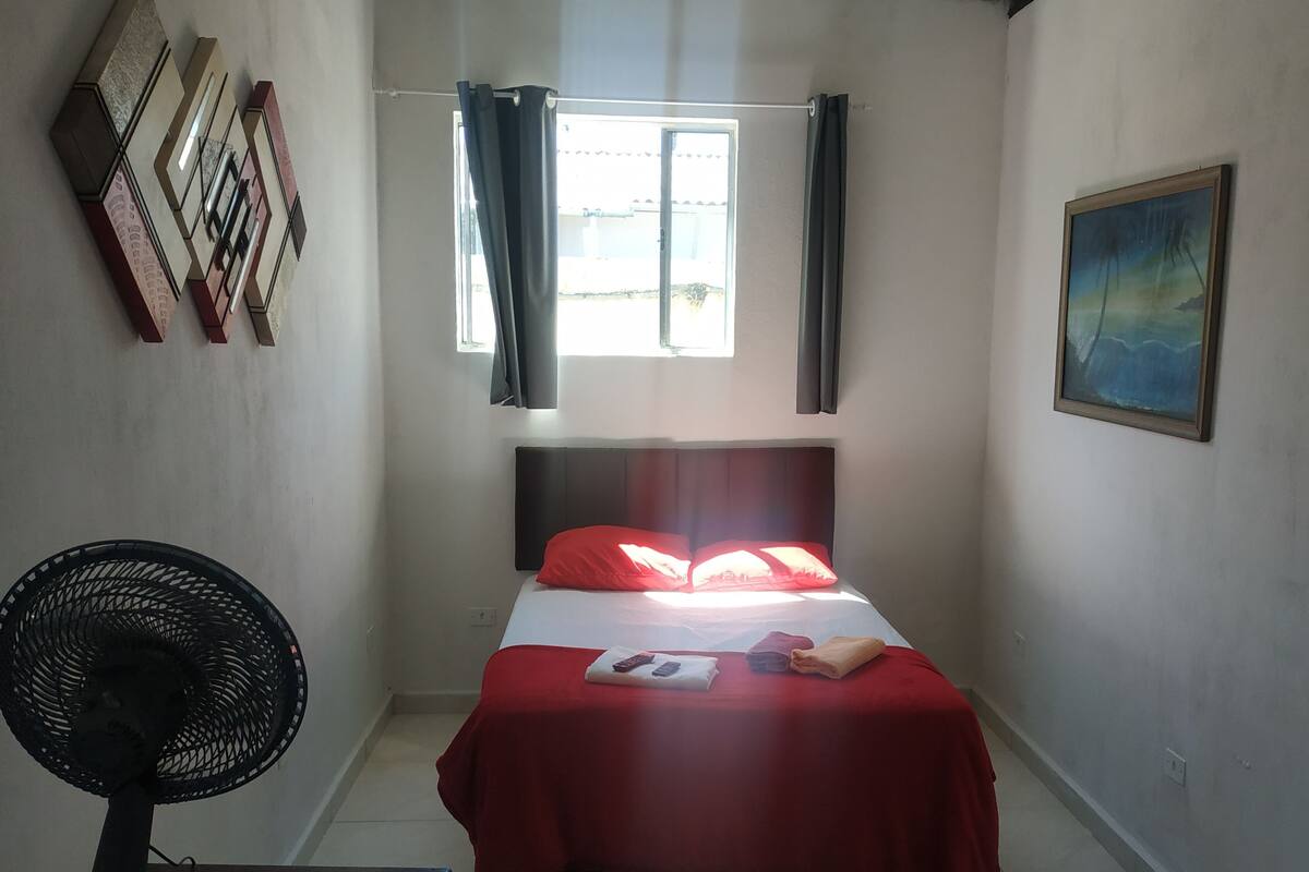 A bedroom with a bed, a fan, a window with curtains, a painting, and a wall-mounted shelf.